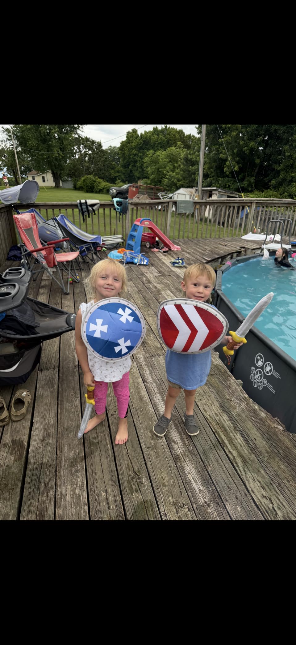 Two young children standing on a wooden deck next to a swimming pool, holding toy shields and a ball. The child on the left holds a blue ball with white stars and a sword, while the child on the right holds a red and white striped shield and a sword.